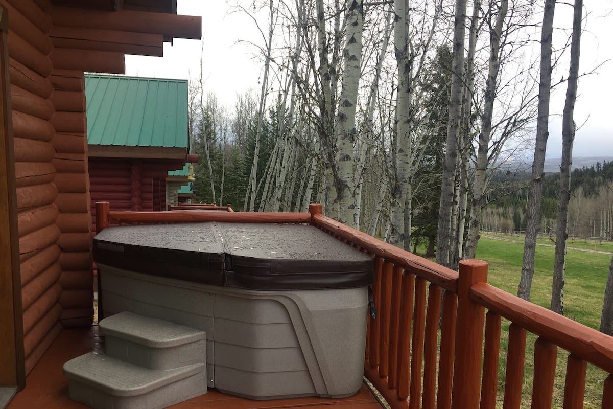 Una terraza de madera con un jacuzzi, escalones que llevan hacia él, y una vista de árboles y un edificio al fondo.