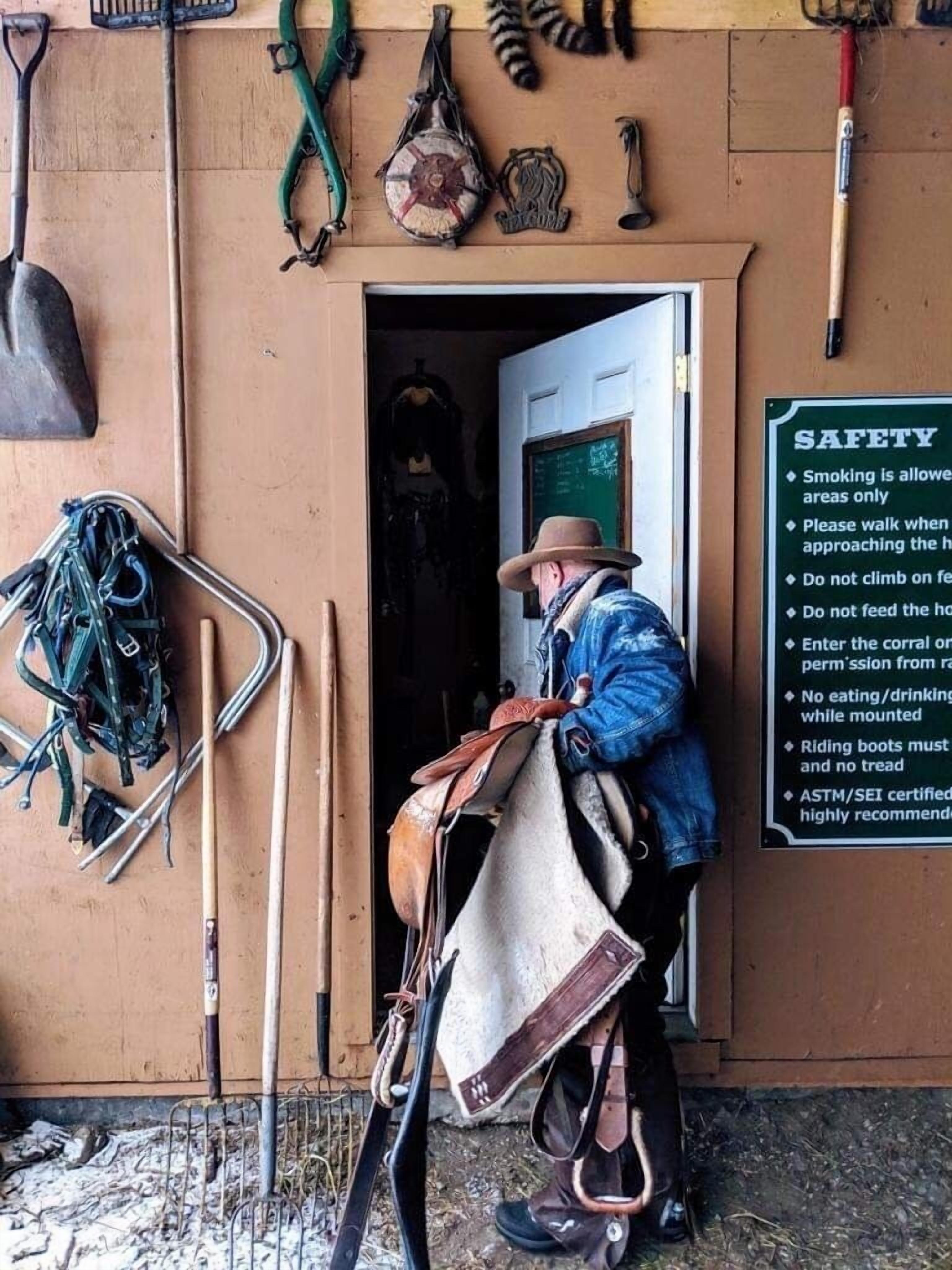 A person in a cowboy hat and denim jacket holding a saddle, standing in front of a door with a safety sign.