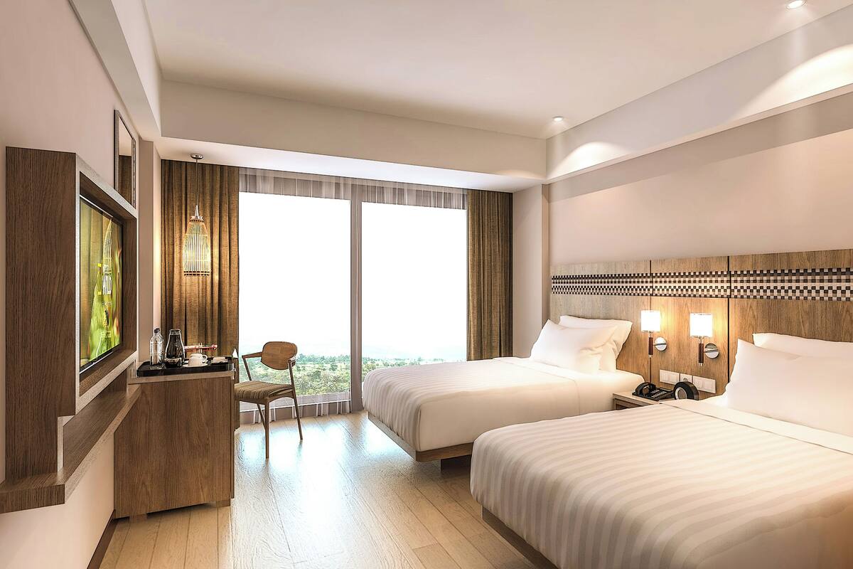 Ein Hotelzimmer mit zwei Betten, einem Schreibtisch mit Stuhl, einem Fernseher und einem großen Fenster mit Vorhängen.