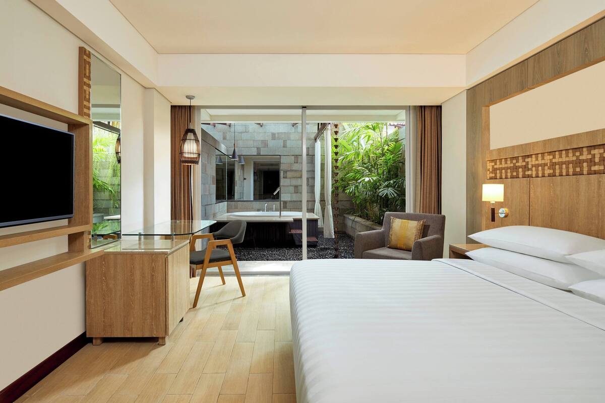 Ein Hotelzimmer mit Bett, Schreibtisch mit Stuhl, Fernseher, Badezimmer mit Waschbecken und Spiegel sowie Blick auf Grünfläche.