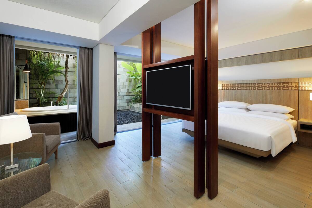 Ein Hotelzimmer mit einem großen Bett, einem Flachbildfernseher, einer Sitzecke mit Tisch und Stühlen sowie Blick auf Grünflächen.
