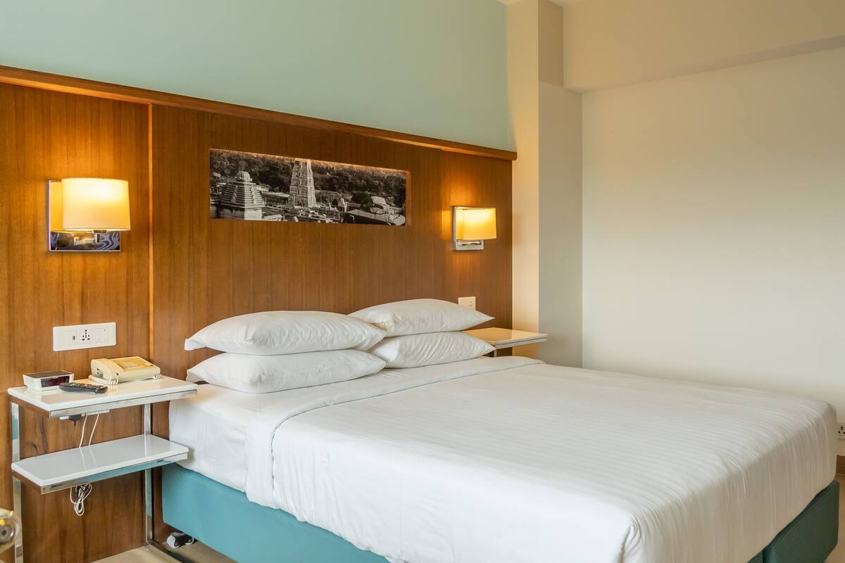 A hotel room with a bed, bedside tables, a nightstand, a wall-mounted lamp, and a picture of a historical building.