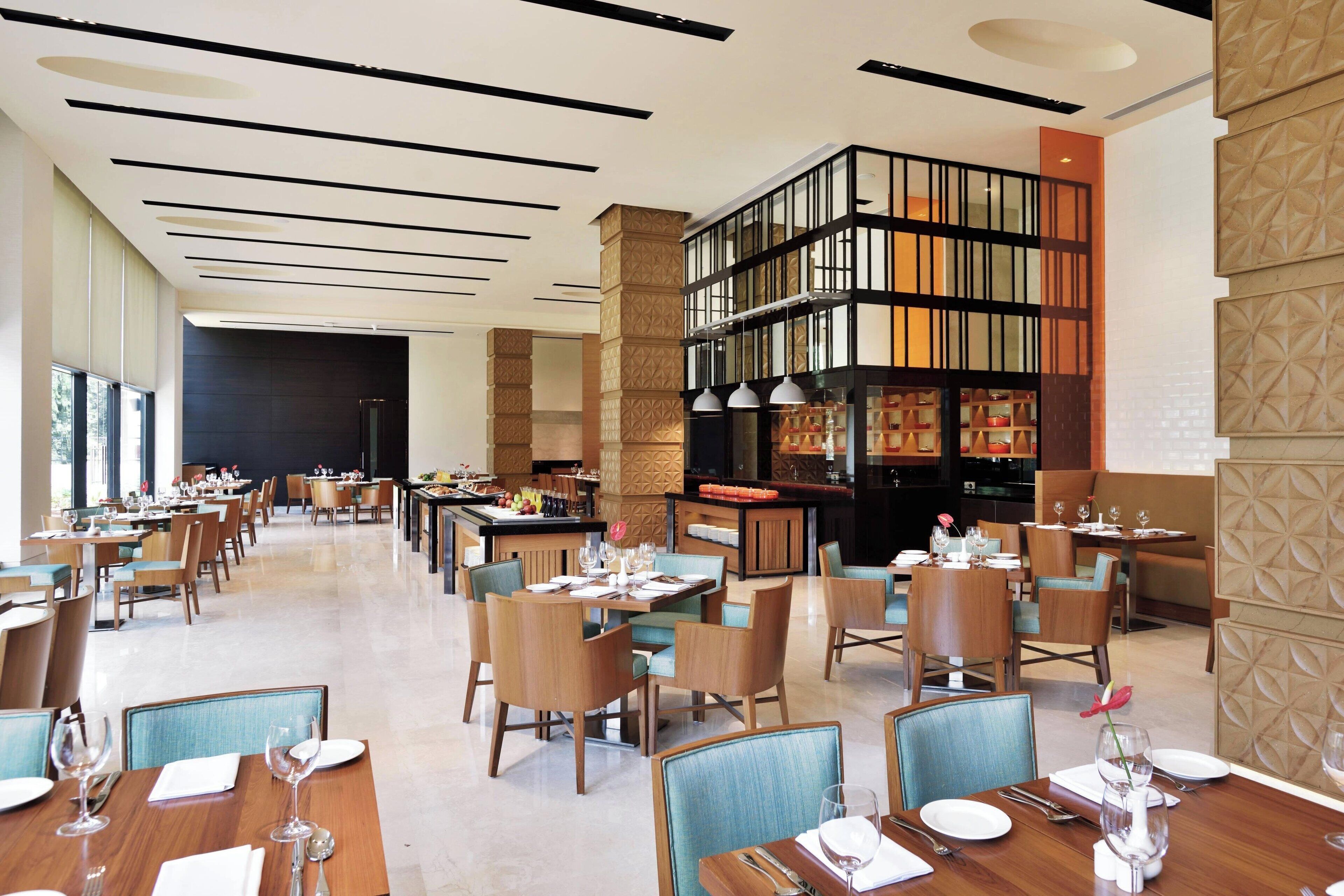 A spacious hotel dining area with multiple tables and chairs, a central bar area, and a decorative bookshelf.