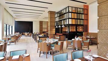 A spacious hotel dining area with multiple tables and chairs, a central bar area, and a decorative bookshelf.