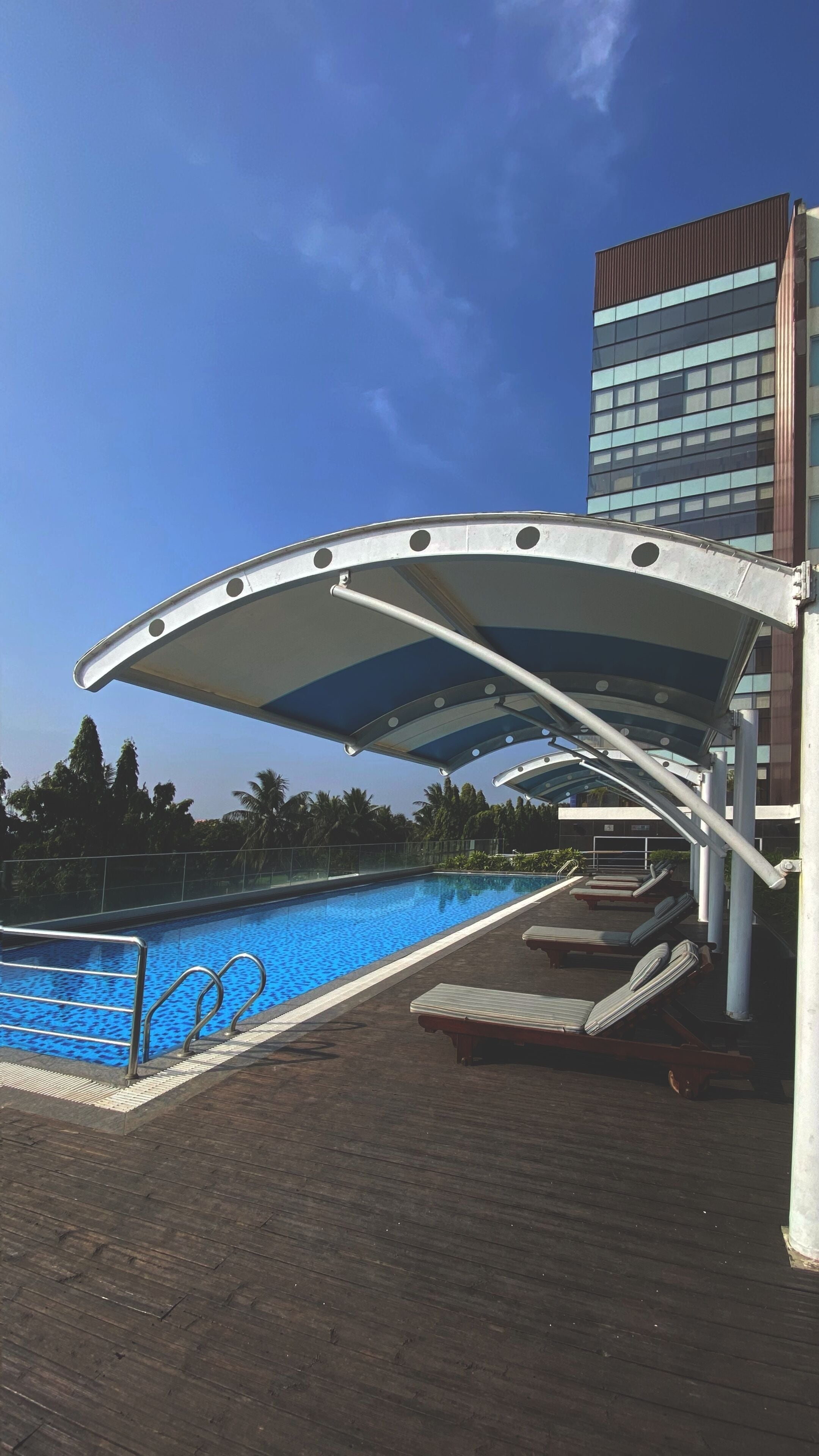Piscina com espreguiçadeiras e área coberta, rodeada por um deck de madeira e um edifício moderno ao fundo.