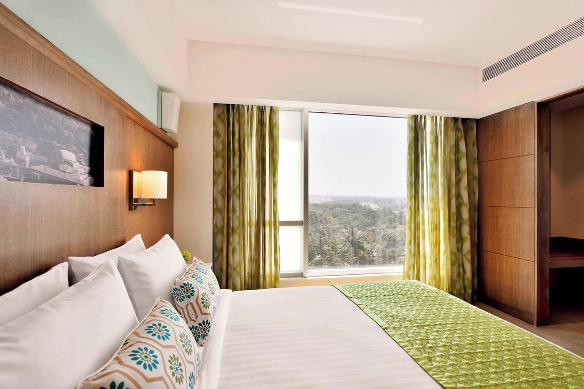 A hotel room with a large bed, a nightstand, a wall-mounted lamp, and a window with curtains.