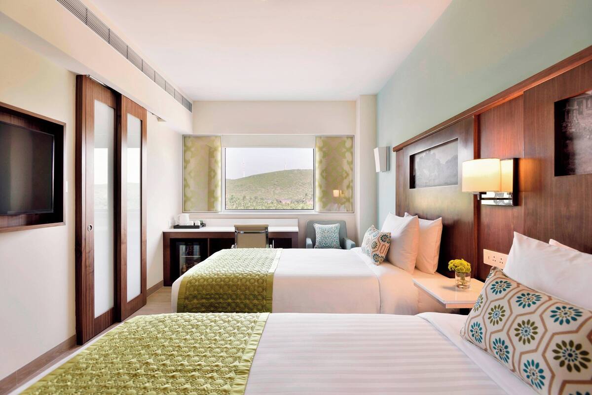A hotel room with two beds, a desk, a chair, a TV, and a window with a view of a hill.