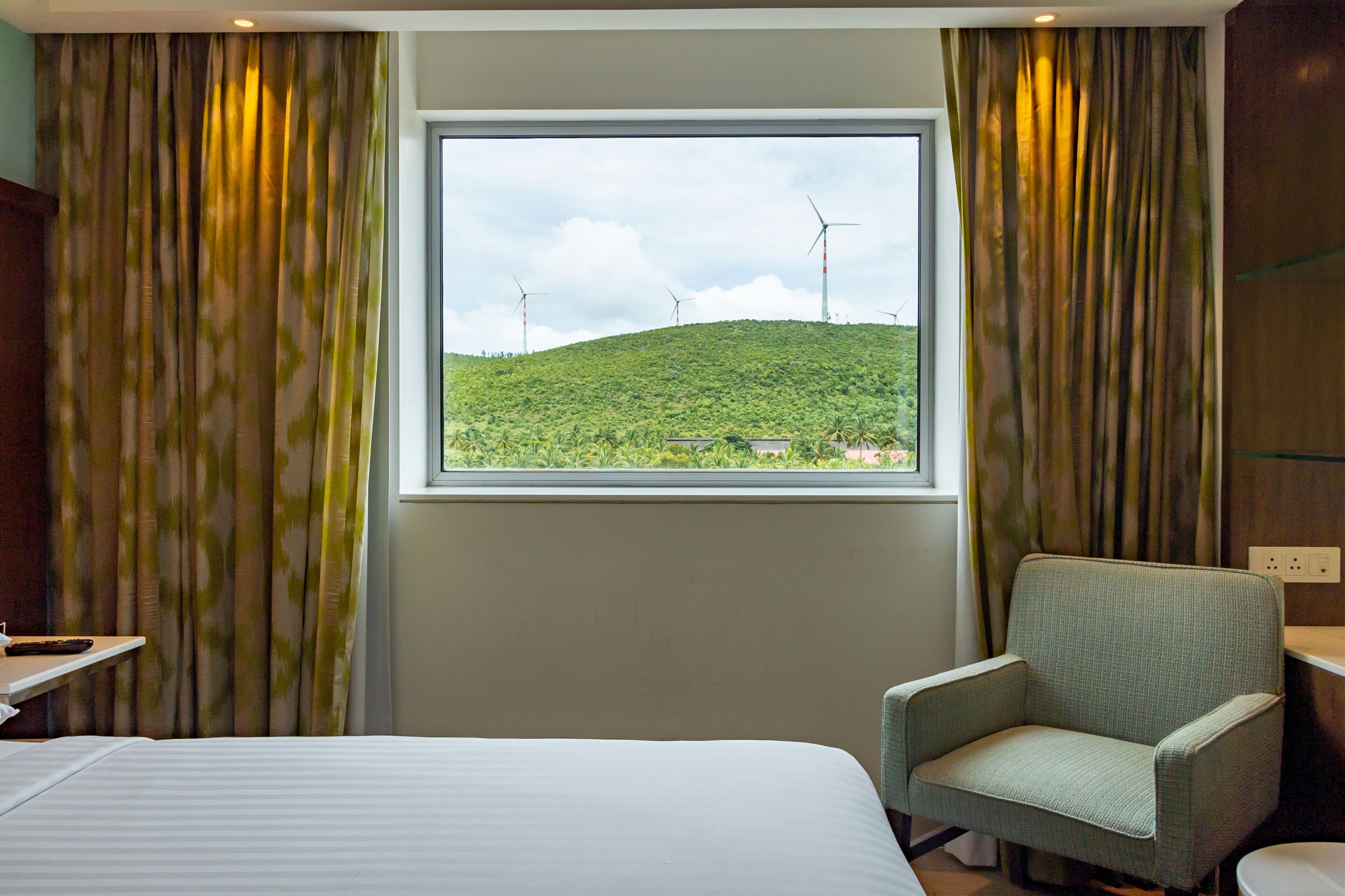 A hotel room with a bed, a chair, a window with a view of wind turbines and greenery.
