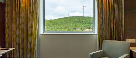 A hotel room with a bed, a chair, a window with a view of wind turbines and greenery.