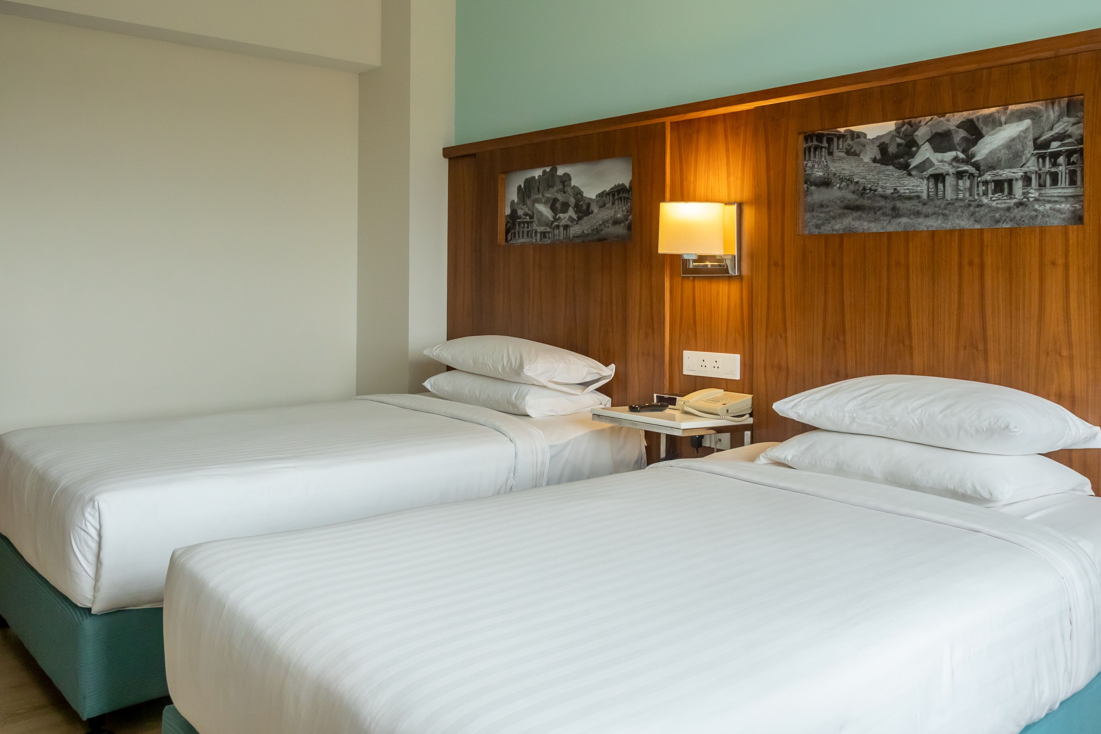 A hotel room with two single beds, a wooden headboard, a nightstand with a lamp, and two wall-mounted paintings.