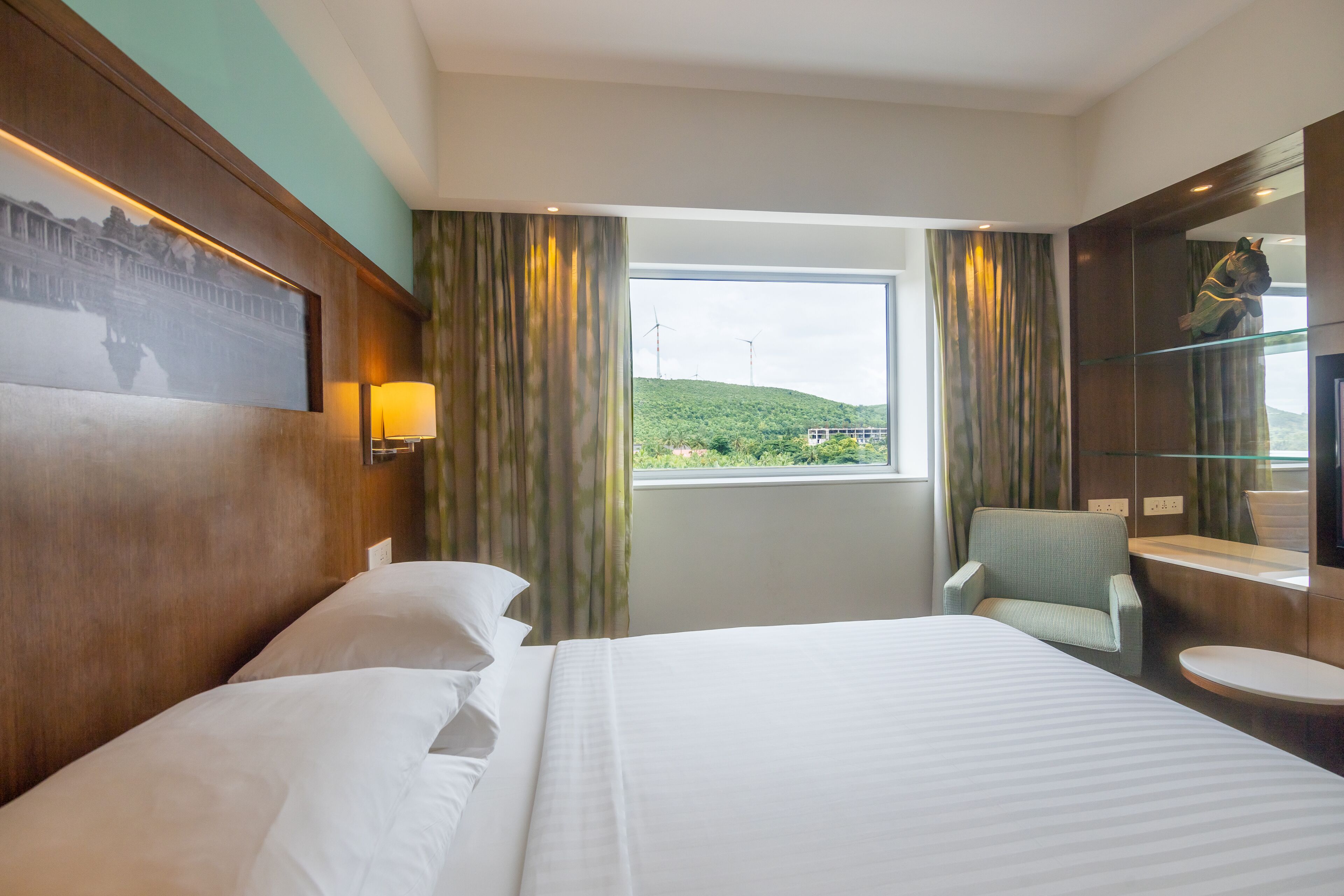 A hotel room with a large bed, a chair, a small table, a mirror, and a window with a view of a green landscape and wind turbines.