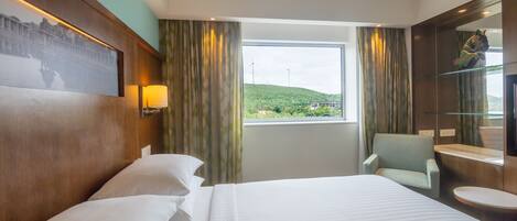 A hotel room with a large bed, a chair, a small table, a mirror, and a window with a view of a green landscape and wind turbines.