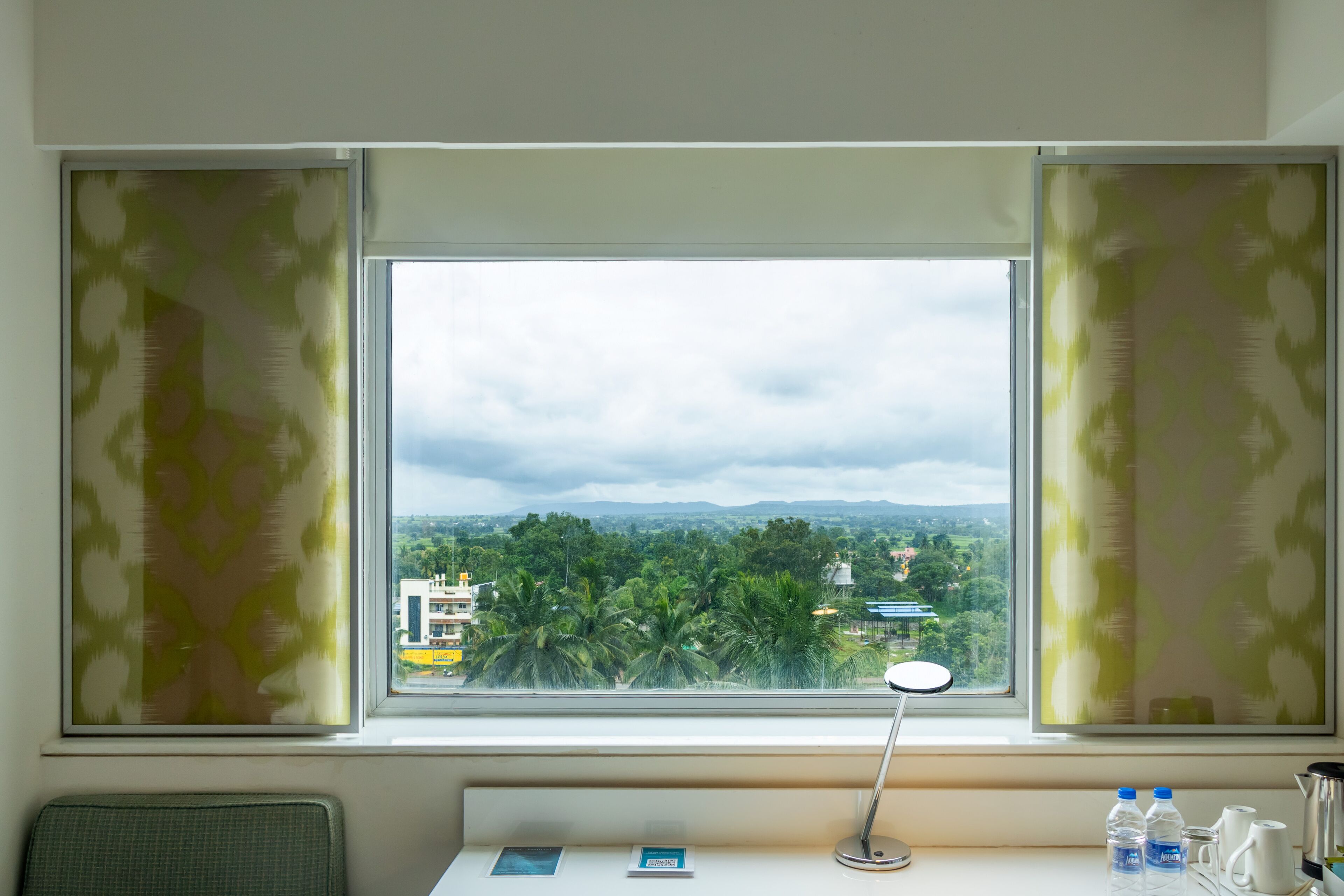 Una habitación con un amplio ventanal, un escritorio con lámpara y vistas a un paisaje urbano con edificios y árboles.