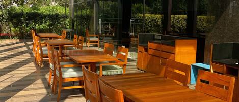 An outdoor seating area with wooden tables and chairs, a glass roof, and a view of greenery.
