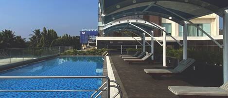 A swimming pool with a wooden deck, lounge chairs, and a covered area.