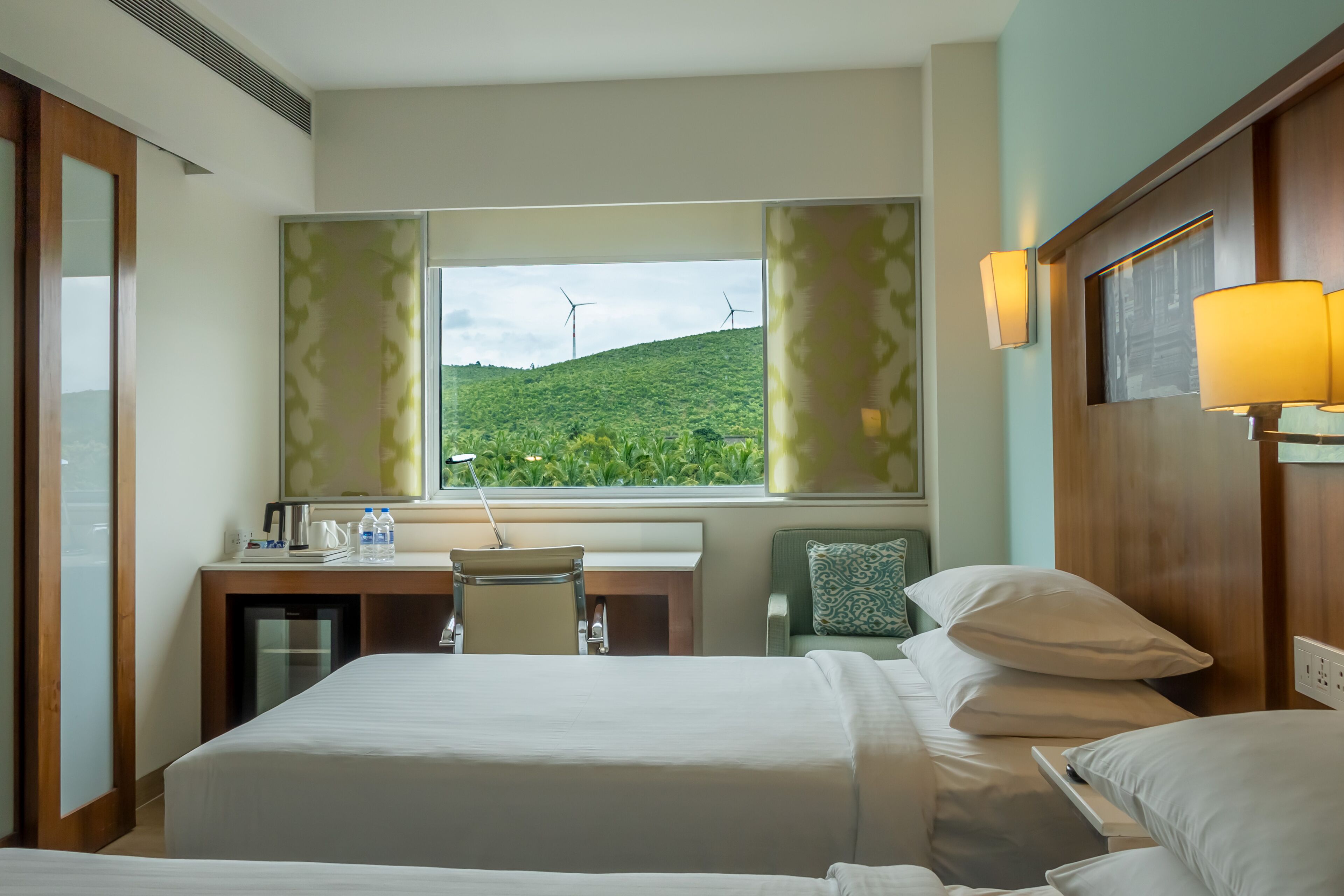 A hotel room with a bed, desk, chair, and a view of wind turbines on a hill.