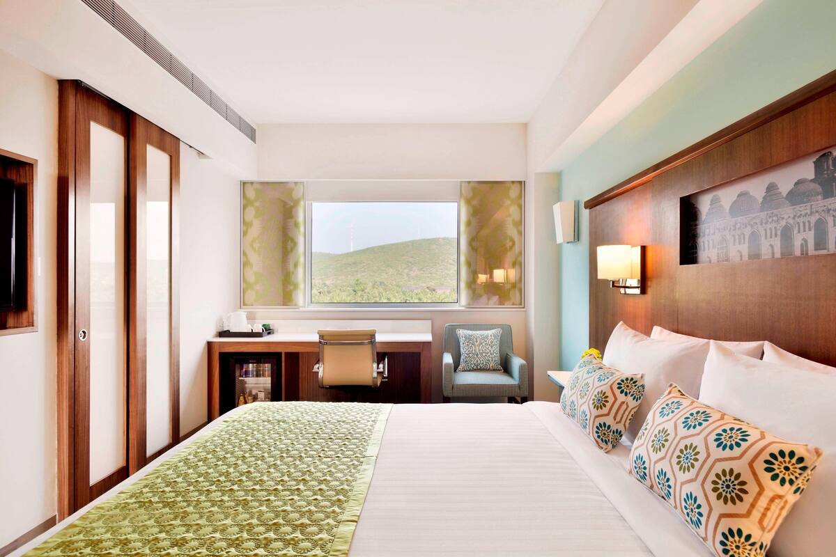 A hotel room with a large bed, a desk, a chair, and a view of a hill.