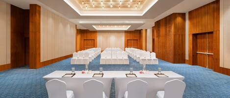 Un salón de conferencias con filas de sillas blancas, mesas con placas de identificación y una alfombra azul estampada.