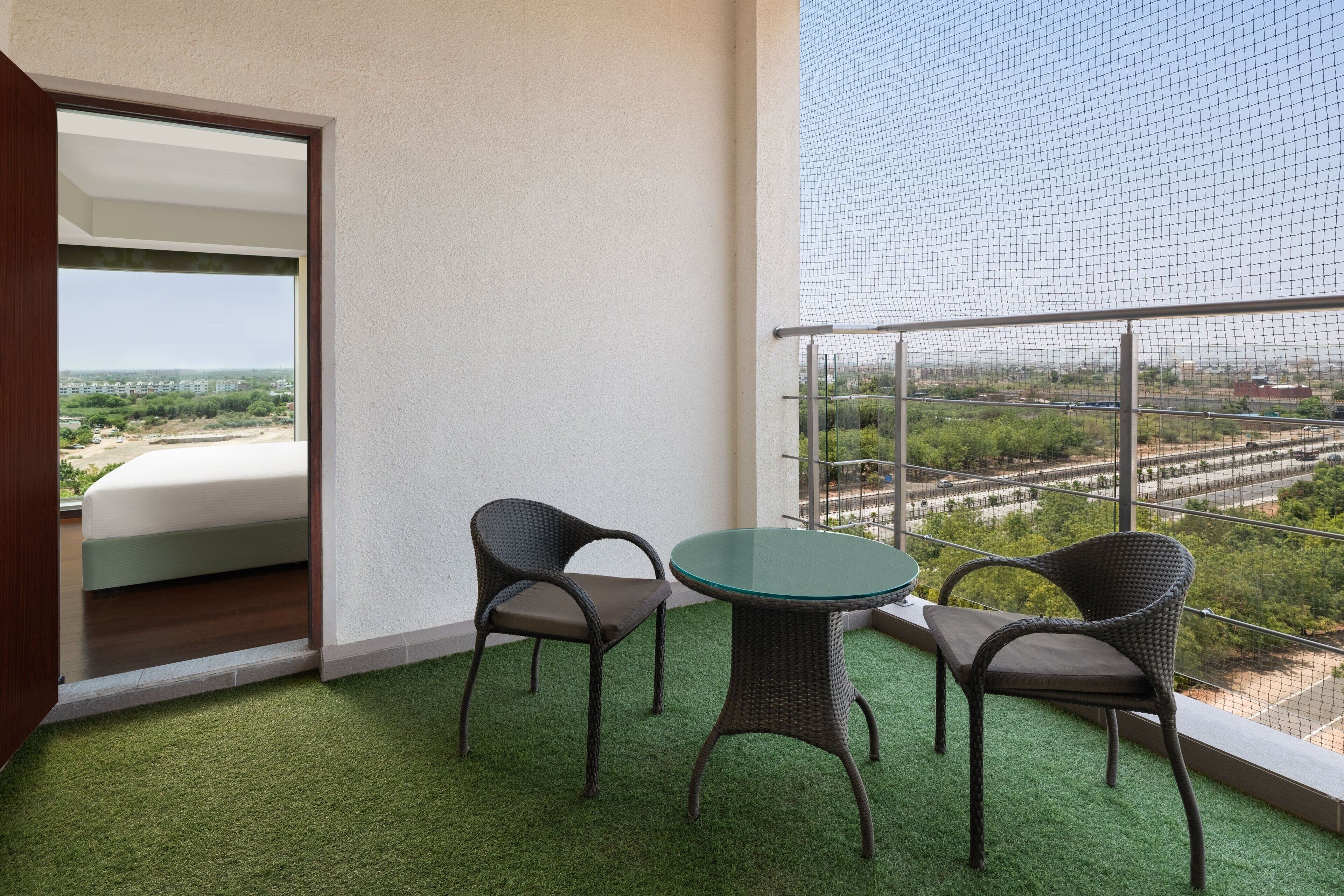 Un balcón con suelo de césped artificial verde, dos sillas de mimbre, una mesa de vidrio y vistas a un paisaje urbano a través de una malla metálica.