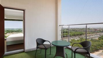 A balcony with a green artificial turf floor, two wicker chairs, a glass table, and a view of a cityscape through a wire mesh.