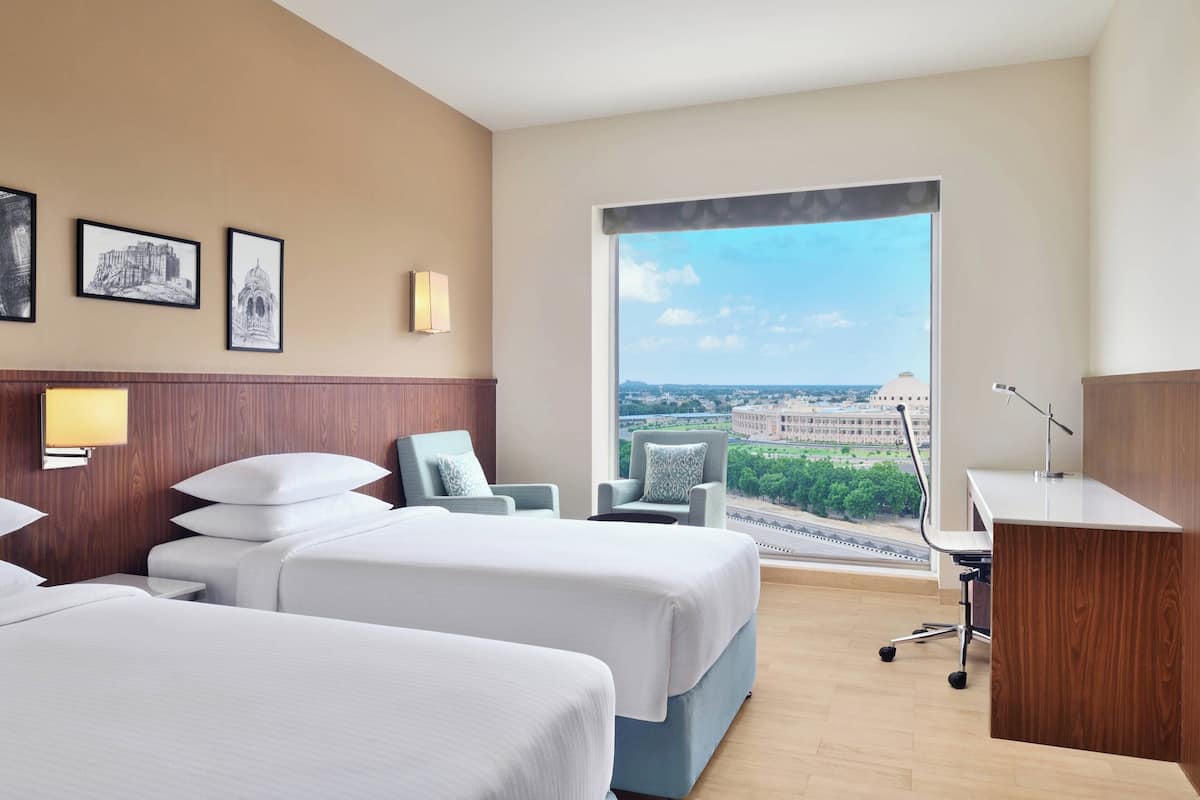 Habitación de hotel con dos camas, un escritorio, una silla y un ventanal con vista a la ciudad.