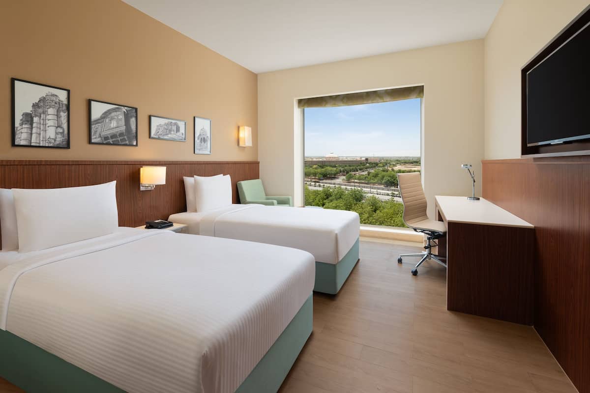 Habitación de hotel con dos camas, un escritorio, una silla y un amplio ventanal con vistas a la ciudad.