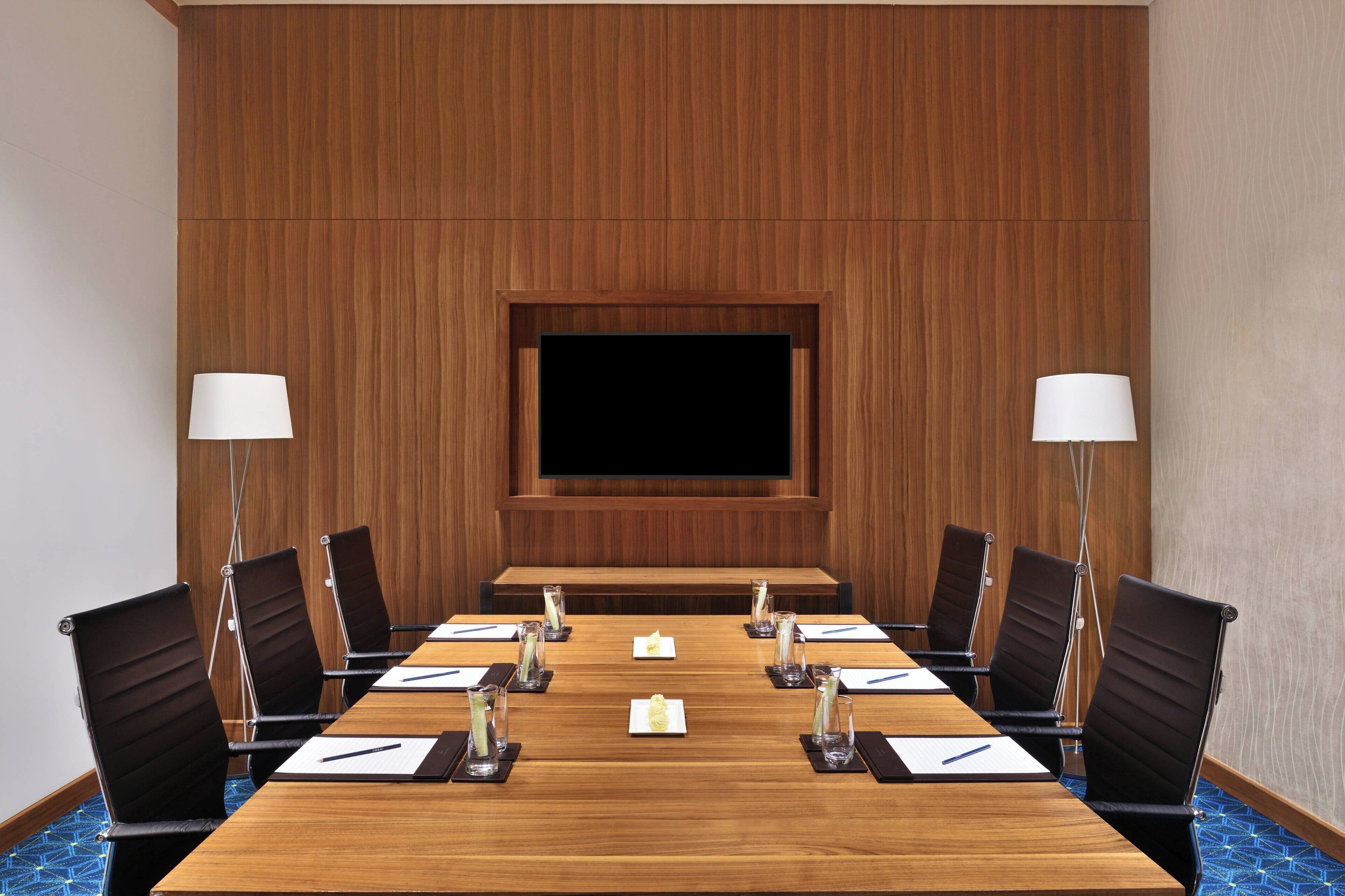 Un salón de conferencias con una mesa de madera, sillas negras, una pantalla grande y dos lámparas.