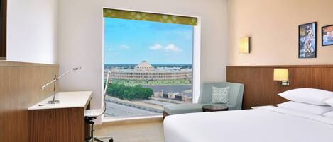 A hotel room with a large window, a desk, a chair, a bed, and a nightstand.