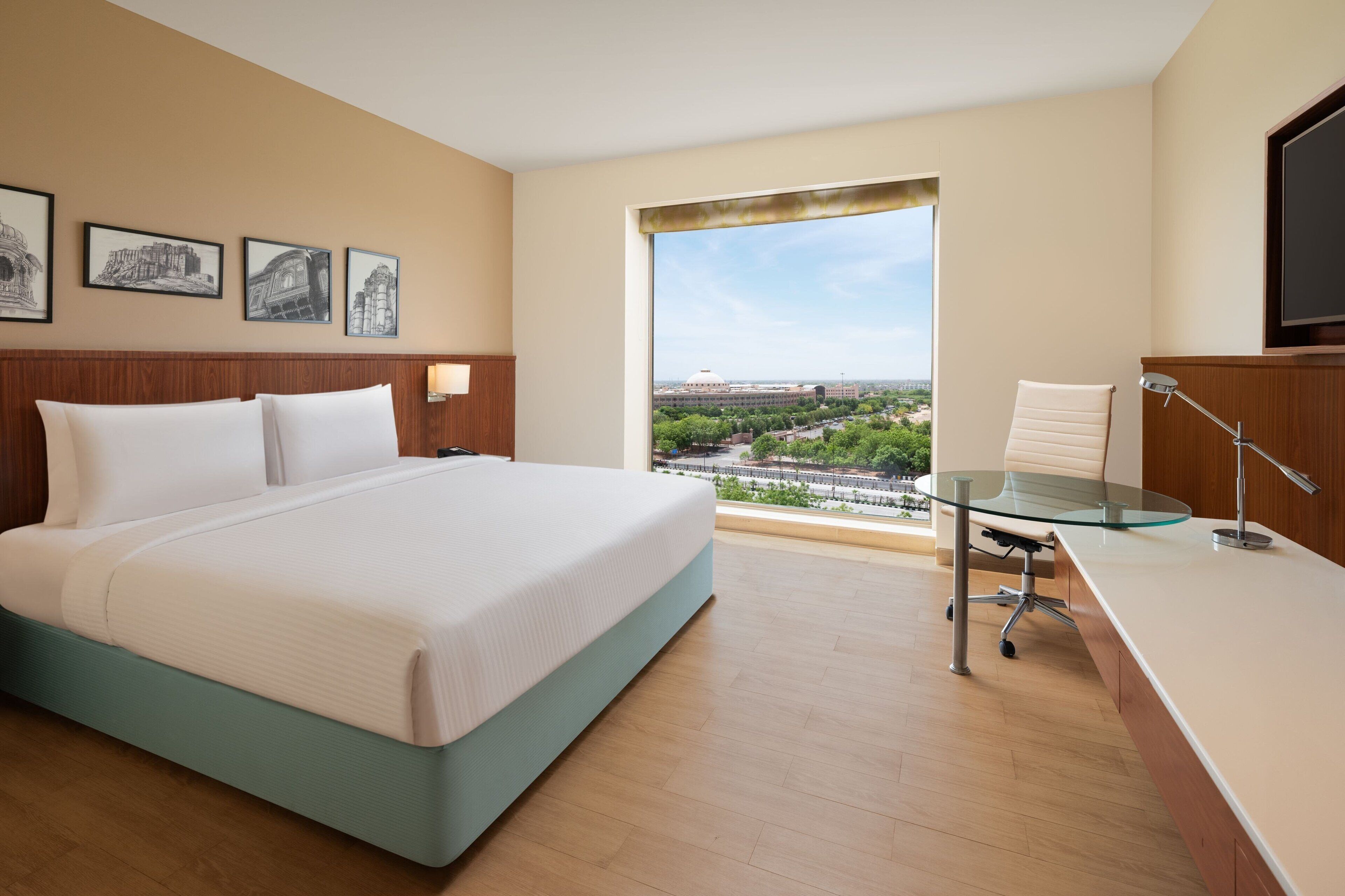 Une chambre d’hôtel avec un grand lit, un bureau doté d’un plateau en verre et une vue sur le paysage urbain, visible à travers une grande fenêtre.
