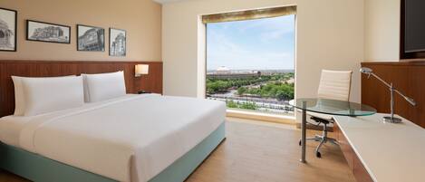 Une chambre d’hôtel avec un grand lit, un bureau doté d’un plateau en verre et une vue sur le paysage urbain, visible à travers une grande fenêtre.