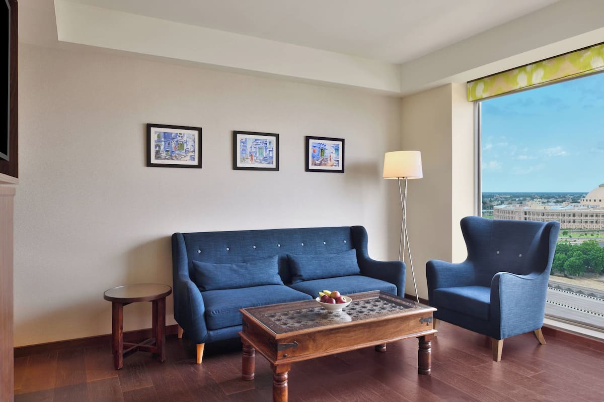 Una habitación con un sofá azul, un sillón a juego, una mesita redonda con un frutero y una lámpara de pie. En la pared hay tres cuadros y una ventana grande con vistas a edificios.