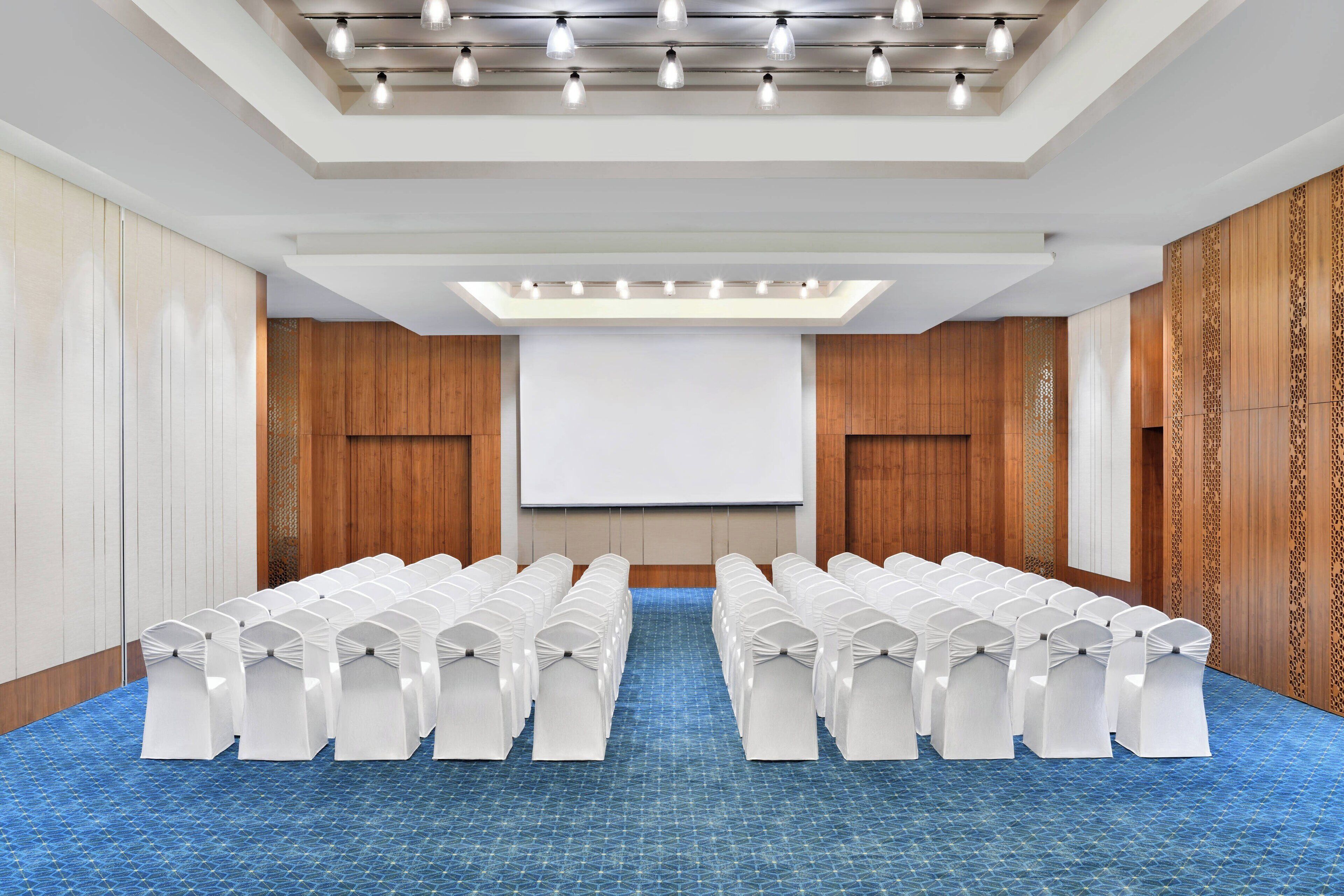 A conference room with rows of white chairs, a projector screen, and a patterned carpet.