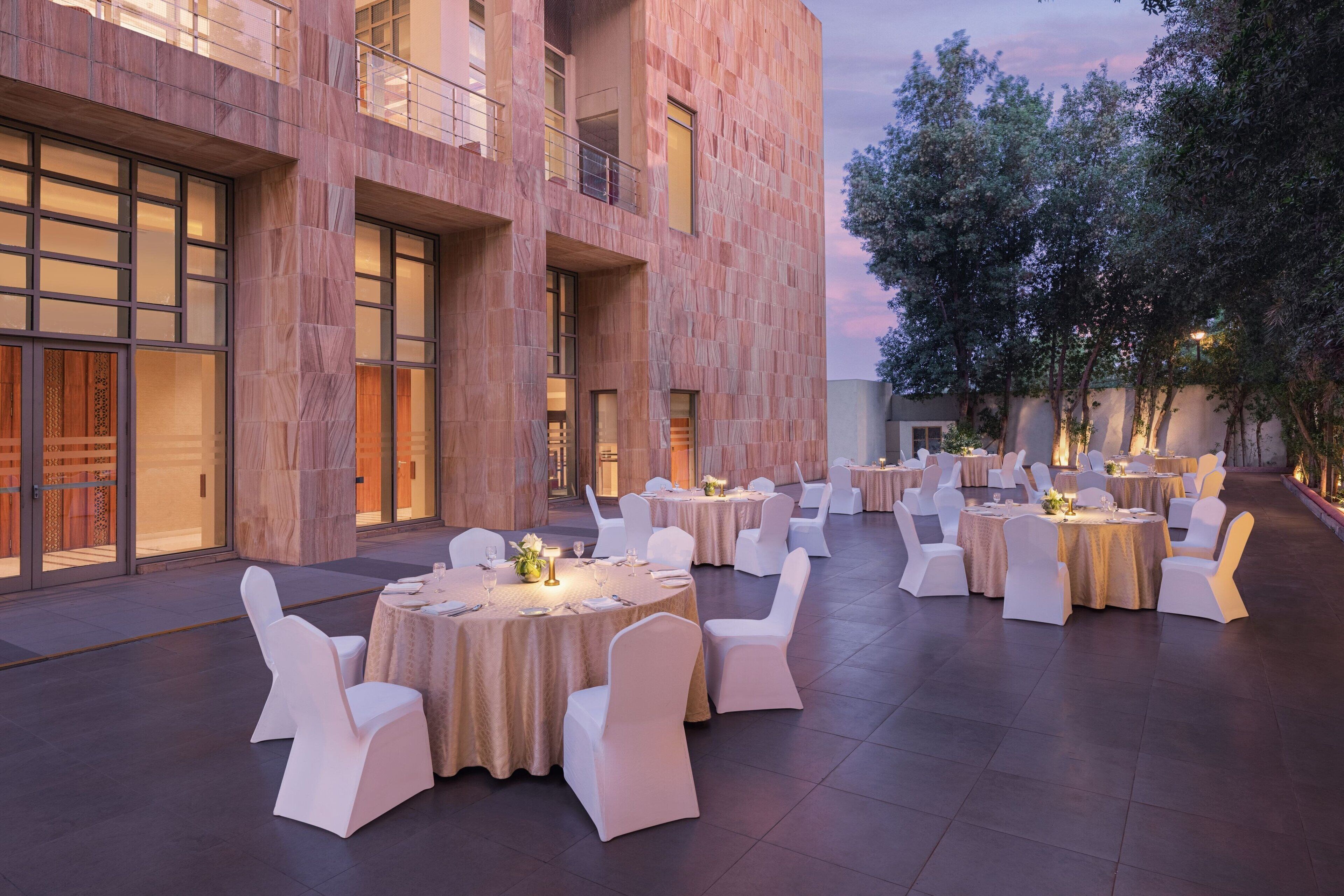 Un espace de restauration en plein air, avec des tables rondes et des chaises blanches, situé devant un bâtiment moderne doté de grandes fenêtres et d’une façade en pierre.