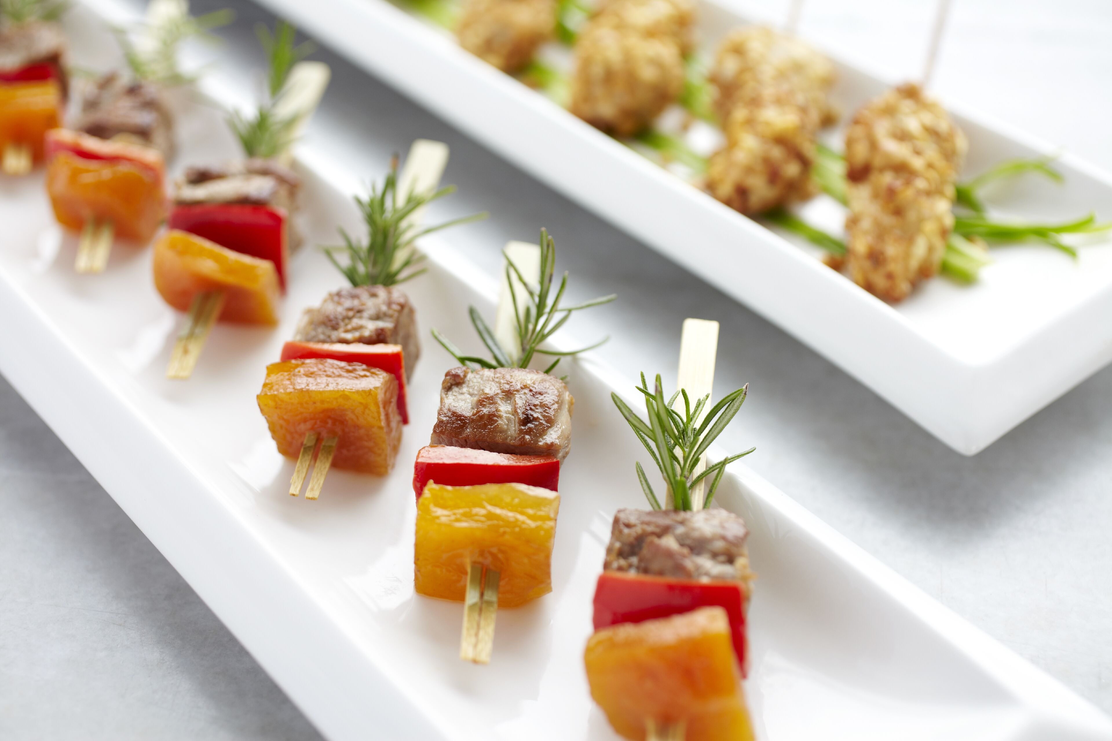 A white rectangular plate with skewers of meat and vegetables, garnished with rosemary.