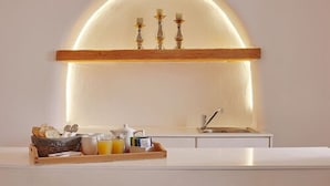 A modern kitchen with a white countertop, a wooden shelf with three candles, and a breakfast tray with bread, eggs, and orange juice. - Ayoba Santorini (Santorini)