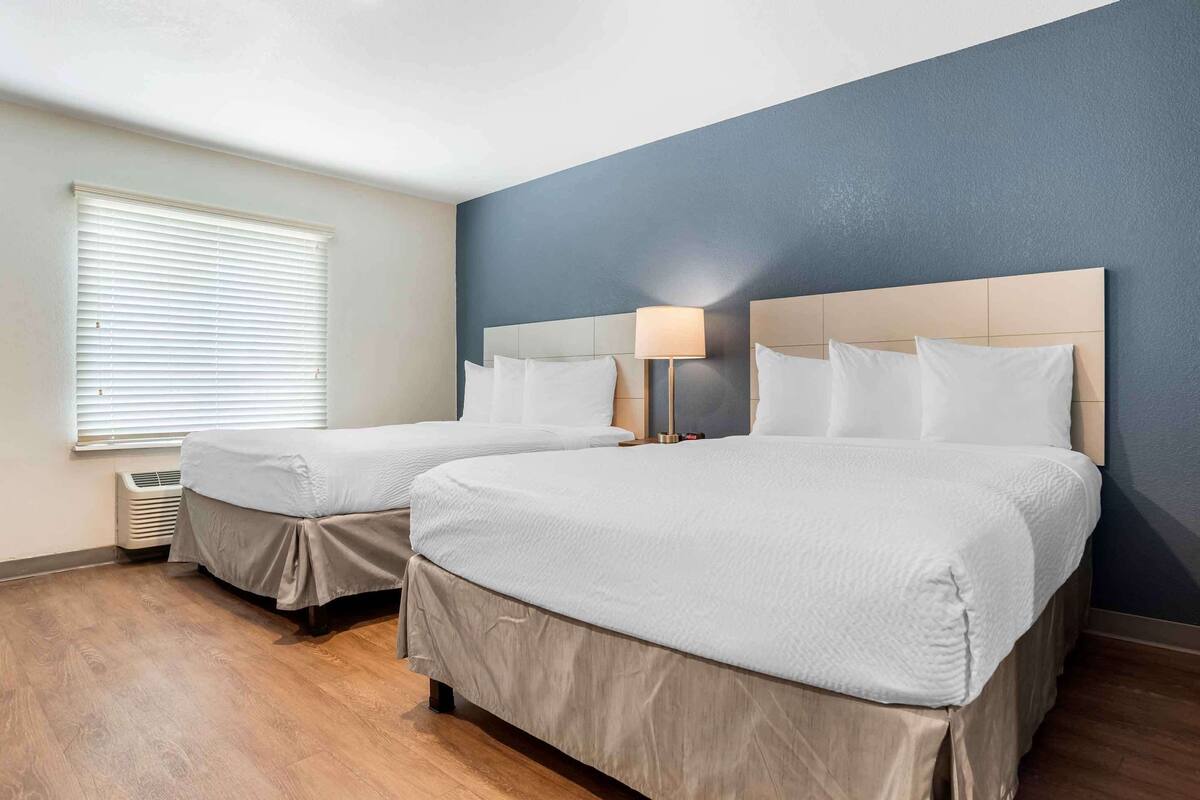 Une chambre d’hôtel avec deux lits, une fenêtre avec des stores, un radiateur mural et une table de chevet avec une lampe.
