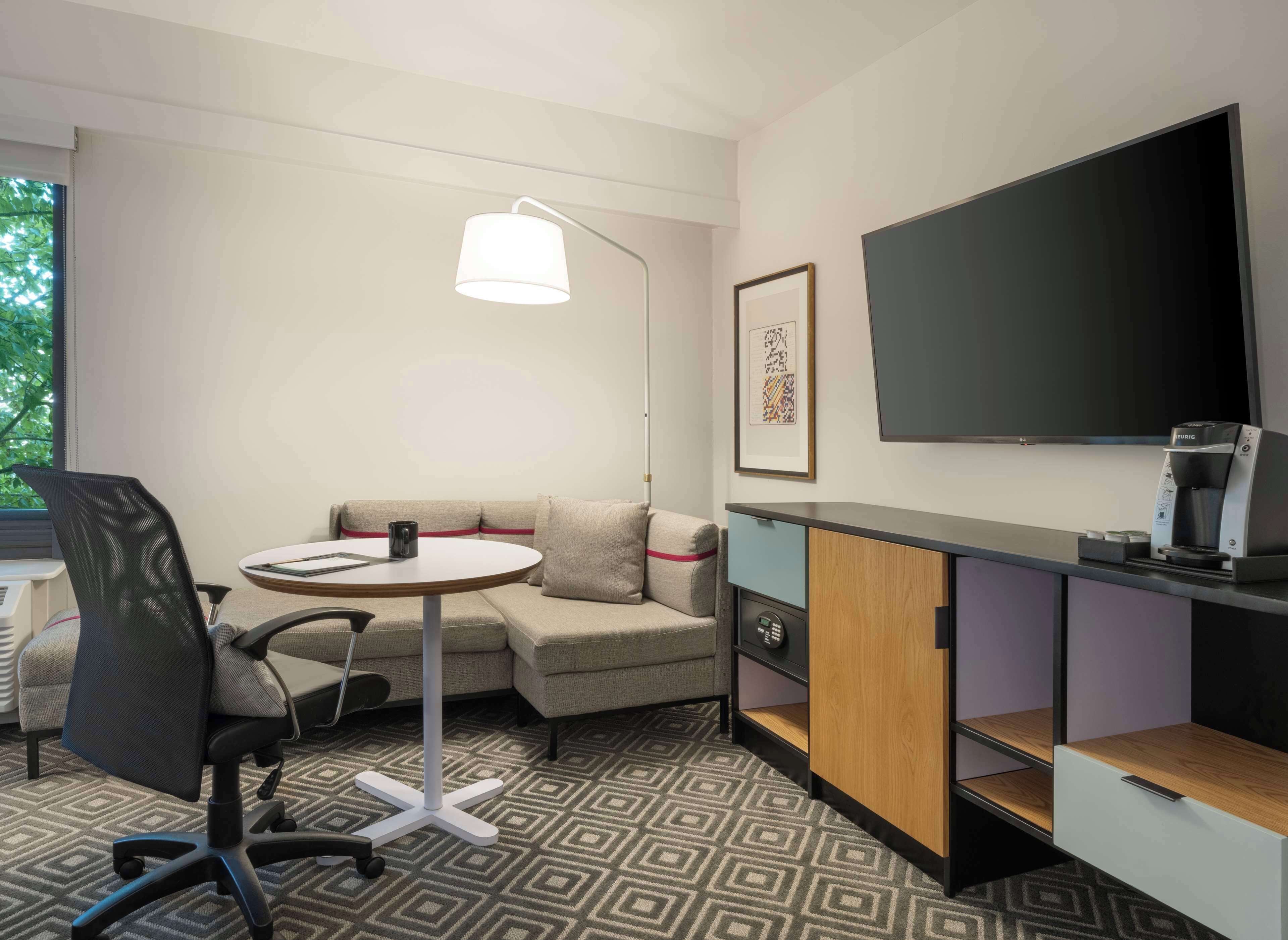 A modern hotel room with a sofa, a round table, a desk with a coffee machine, a TV, and a patterned carpet.