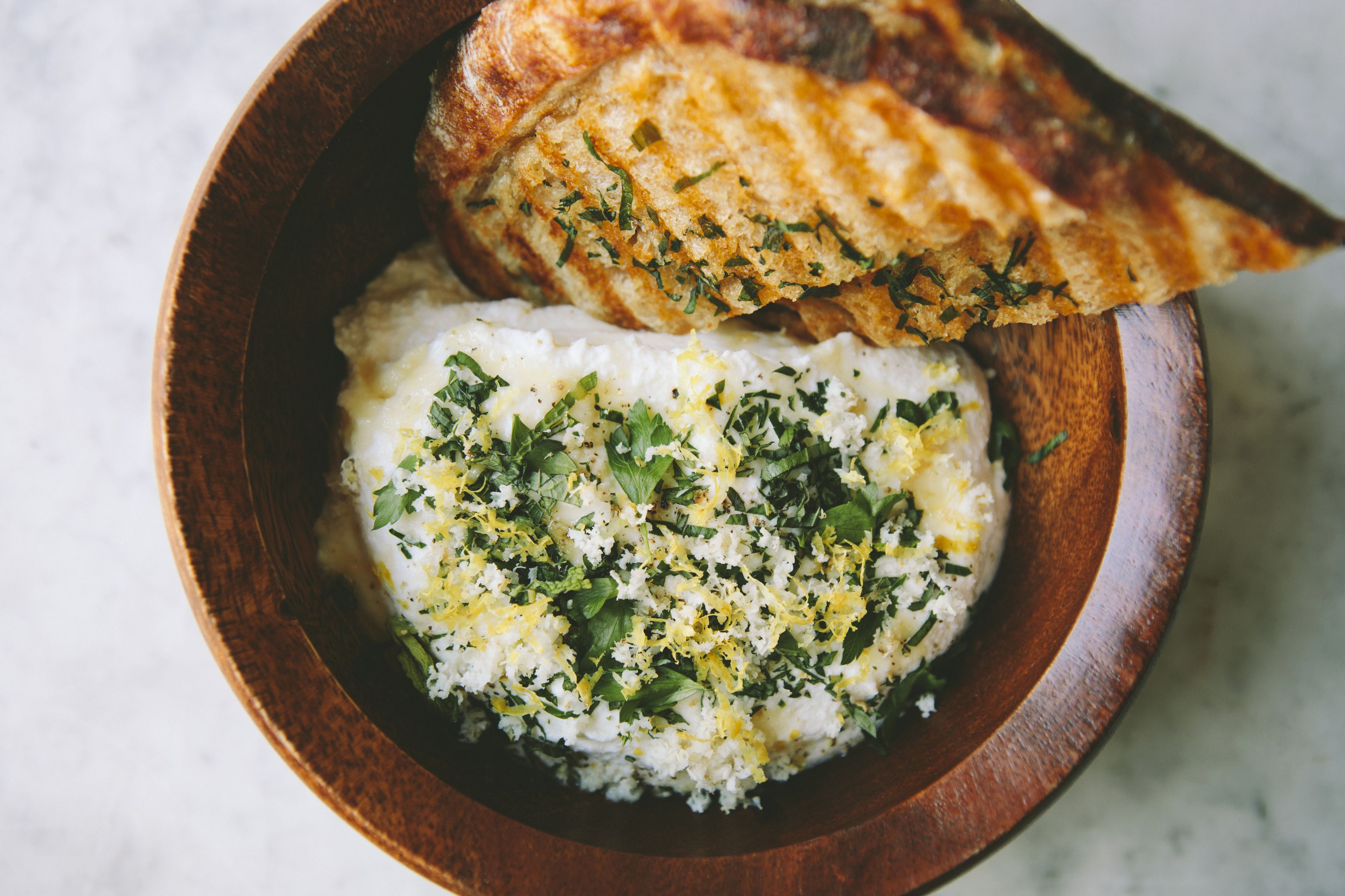 Un bol de fusta amb un plat blanc i cremós, decorat amb herbes i ratlladura de llimona, acompanyat d'una torrada torrada.