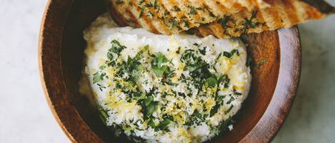 A wooden bowl containing a white creamy dish garnished with herbs and lemon zest, accompanied by a piece of grilled bread.