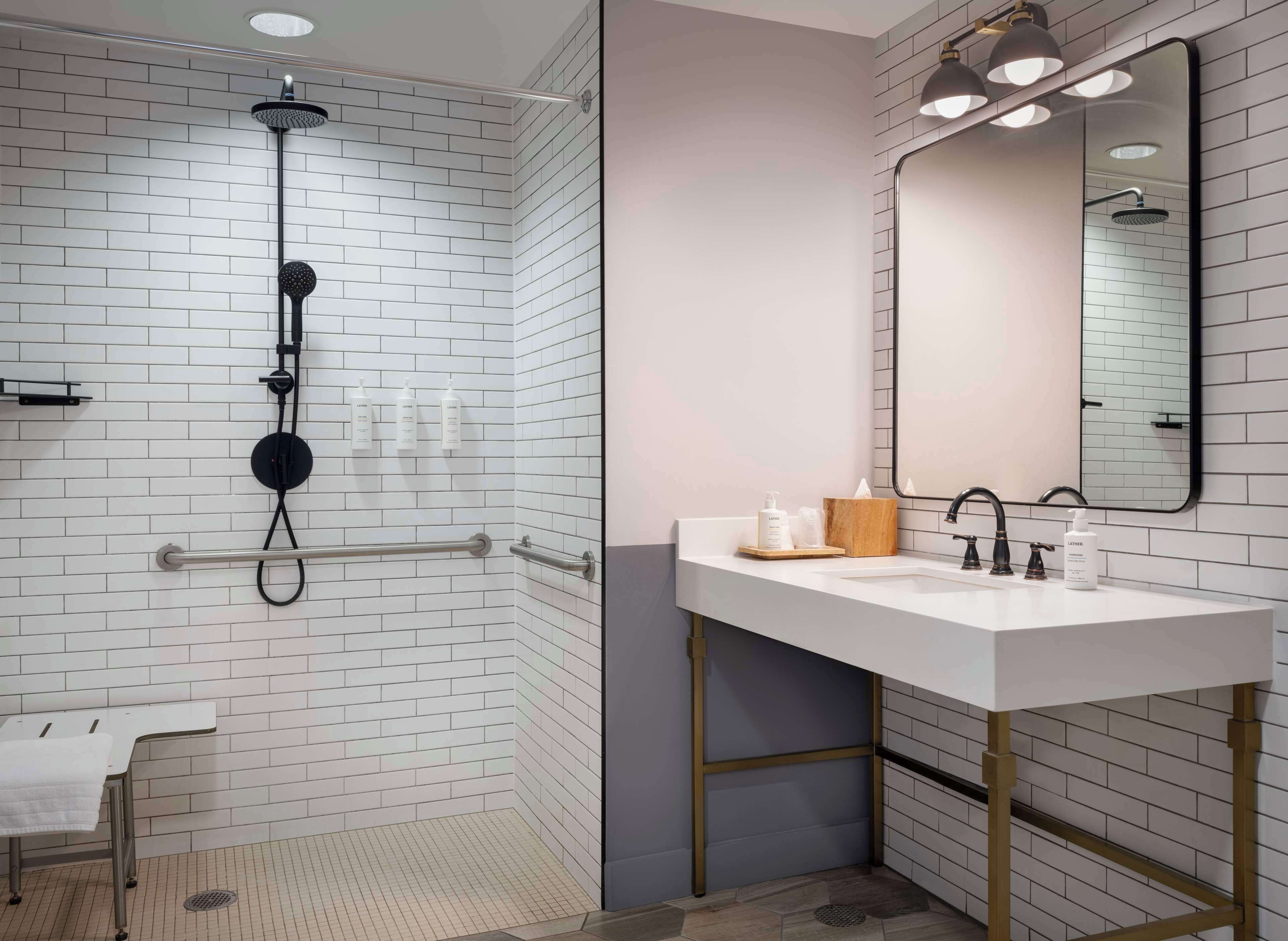 Un baño moderno con ducha de azulejos blancos, lavamanos blanco y detalles dorados, y un espejo grande.