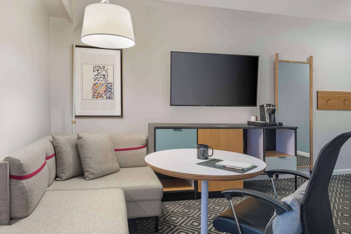 A modern hotel room with a sofa, a round table, a coffee maker, a TV, and a framed artwork.