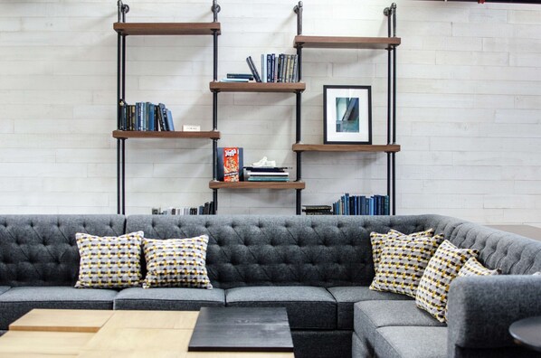 A modern living room with a grey sofa, patterned cushions, and a bookshelf against a white brick wall.