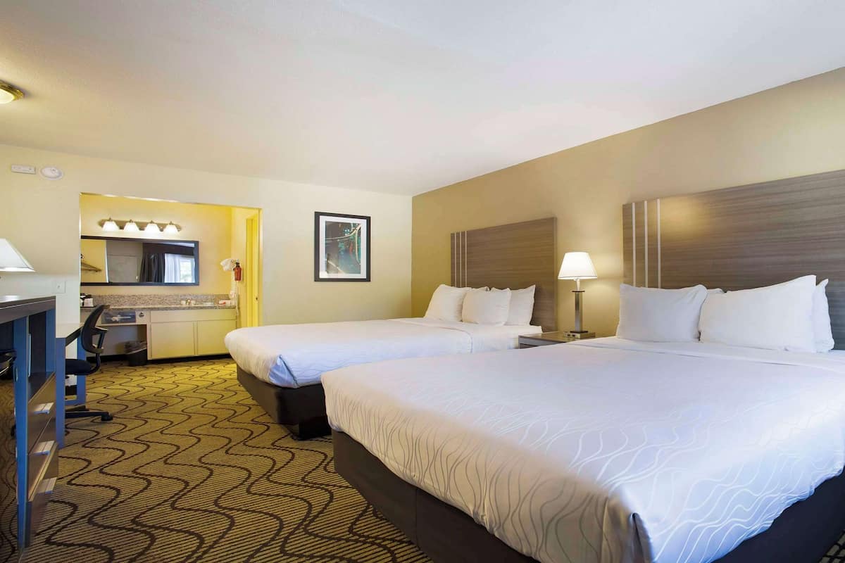 A hotel room with two beds, a desk, a chair, and a bathroom with a mirror and sink.