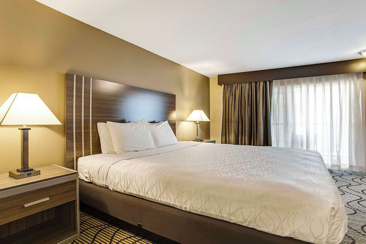 A hotel room with a large bed, two bedside tables with lamps, a headboard, and a window with curtains.
