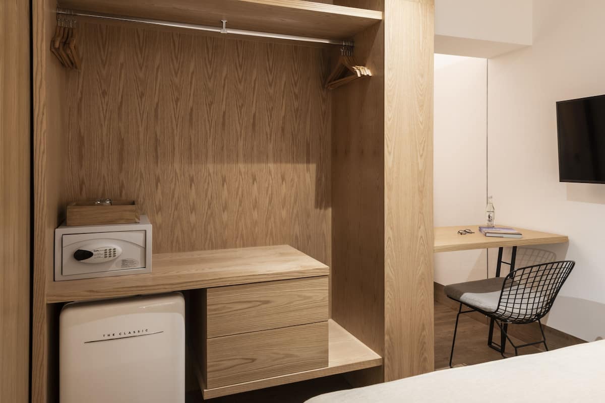 A compact hotel room with a wooden wardrobe, a small desk with a chair, a bed, and a television mounted on the wall.