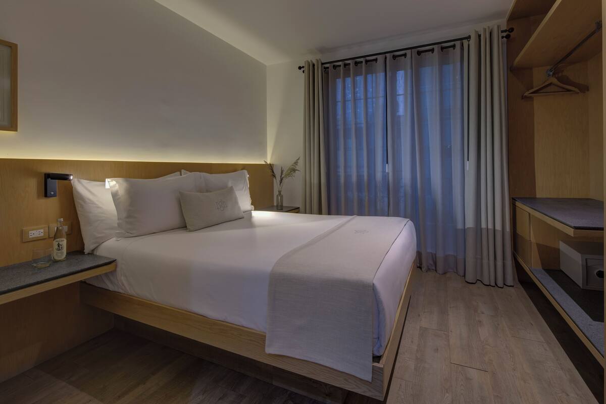 A neatly made bed with a headboard, a bedside table with a bottle, and a hanging coat rack.