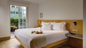 Een groot bed met houten hoofdeinde, een nachtkastje en uitzicht op een balkon met planten.