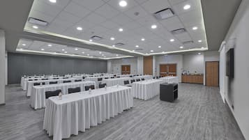 Un amplio salón de conferencias con varias mesas, un escenario y una zona de kitchenette.