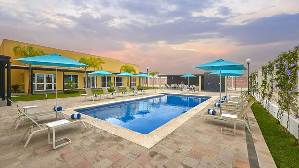 A swimming pool with lounge chairs and umbrellas, surrounded by a building with a yellow facade and a garden. - Hotel Best Place Express (Zamora)