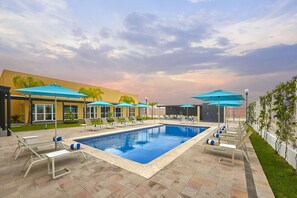 A swimming pool with lounge chairs and umbrellas, surrounded by a building with a yellow facade and a garden. - Hotel Best Place Express (Zamora)